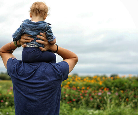 Photo of toddler boy riding on his dad's shoulldlers toward an open field of wildflowrs.